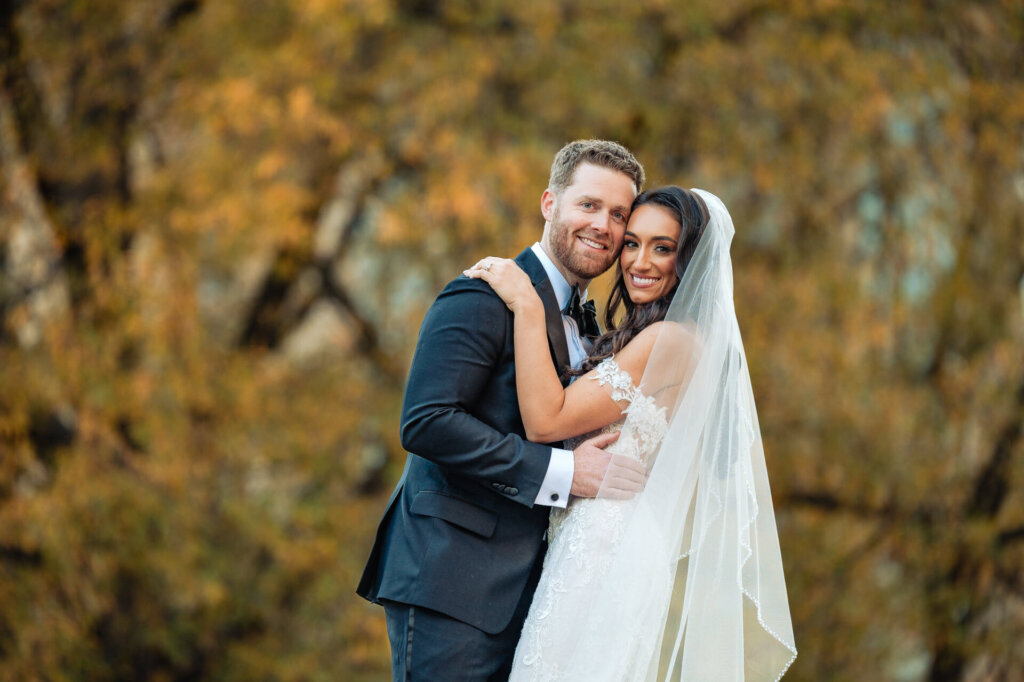Groom embracing the bride from behind as they both smile, framed by golden autumn trees in a park.