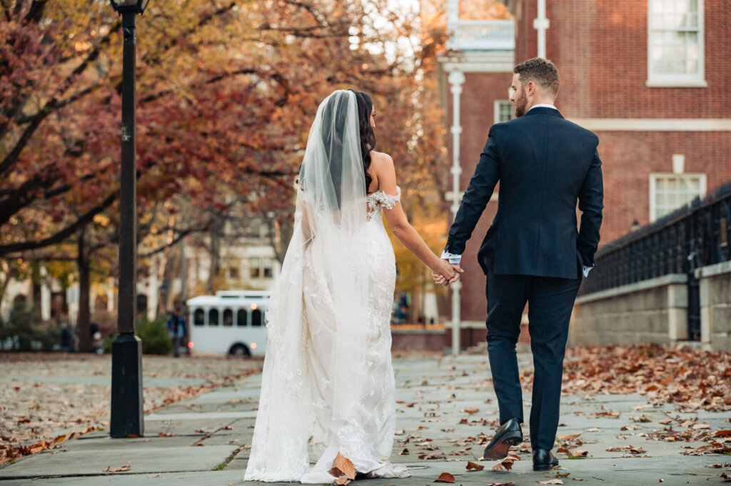 Bride and groom holding hands while walking down a tree-lined sidewalk in an urban setting with autumn leaves scattered around