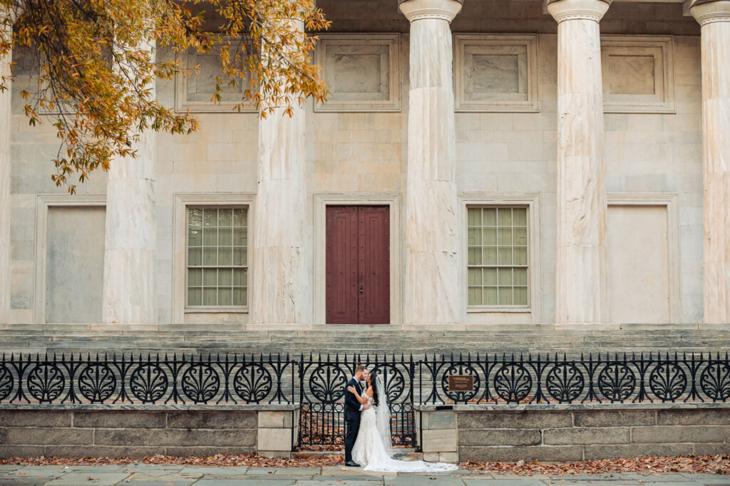 Bride and groom standing together in front of a grand stone building with tall columns, their wedding attire contrasting beautifully with the historic architecture.