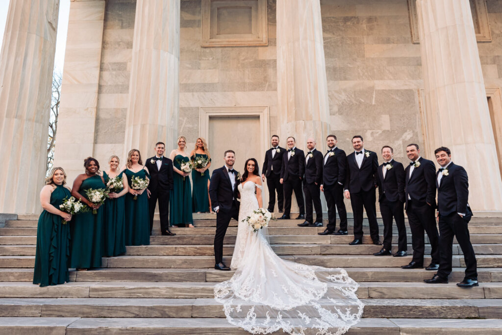 Full bridal party standing on the steps of a grand stone building, with the bride and groom centered and the group wearing green and black formal attire.