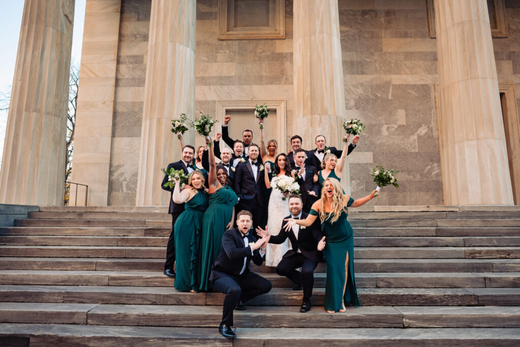 Bridal party and groomsmen playfully jumping and cheering, creating a lively and fun group photo on the steps of a historic building.