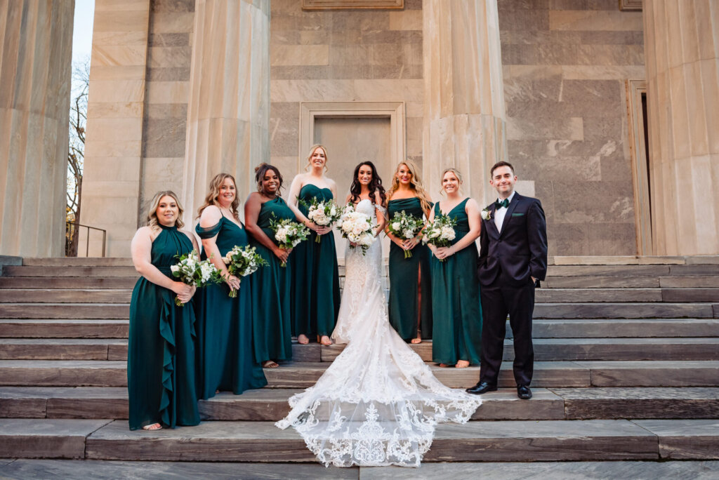 Bride posing with her bridesmaids, all dressed in emerald green gowns, standing on the stone steps of a grand building.