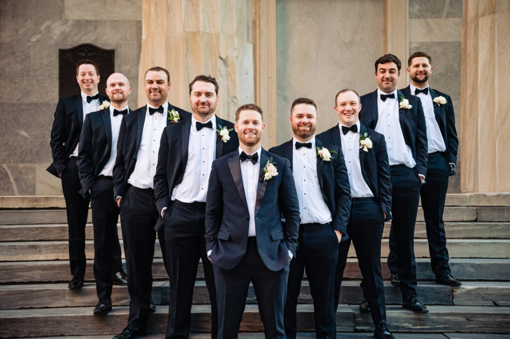Groomsmen standing on the steps of a historic building, dressed in matching tuxedos with boutonnieres, posing confidently as a group.