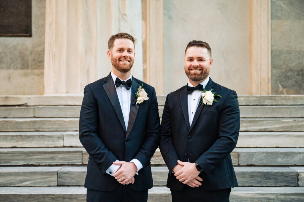Groom posing with his best man on the stone steps of a historic building, both dressed sharply in black tuxedos with matching boutonnieres.
