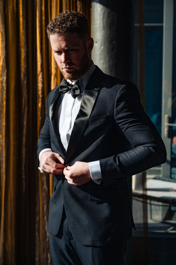 Groom adjusting his tuxedo jacket, standing confidently in a room with elegant golden drapery.