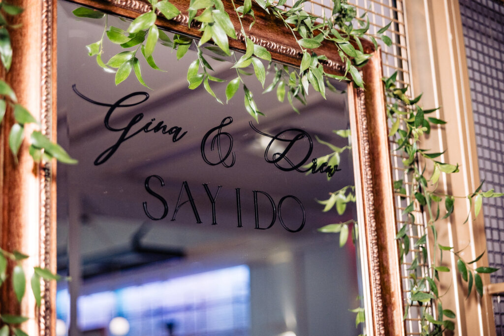 Golden-framed mirror with 'Gina & Drew Say I Do' written in elegant script, adorned with cascading greenery.
