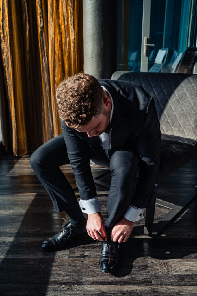 Groom sitting on a chair, bending down to tie his dress shoes in a room with golden curtains and natural light.
