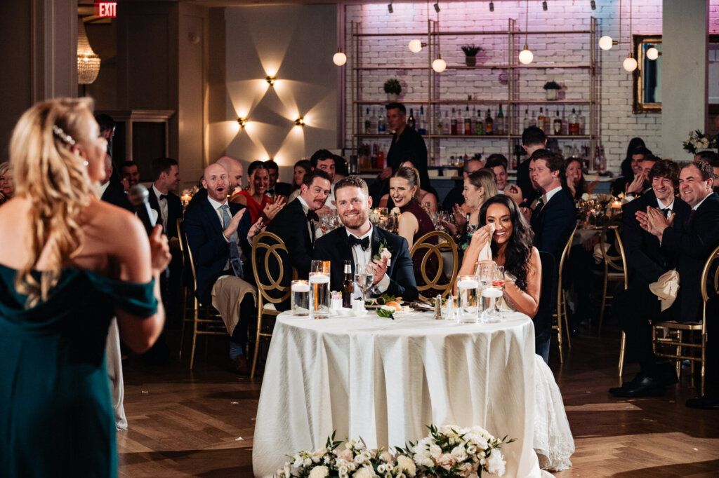 The bridesmaid in an elegant dress giving a speech while standing in front of the seated couple.