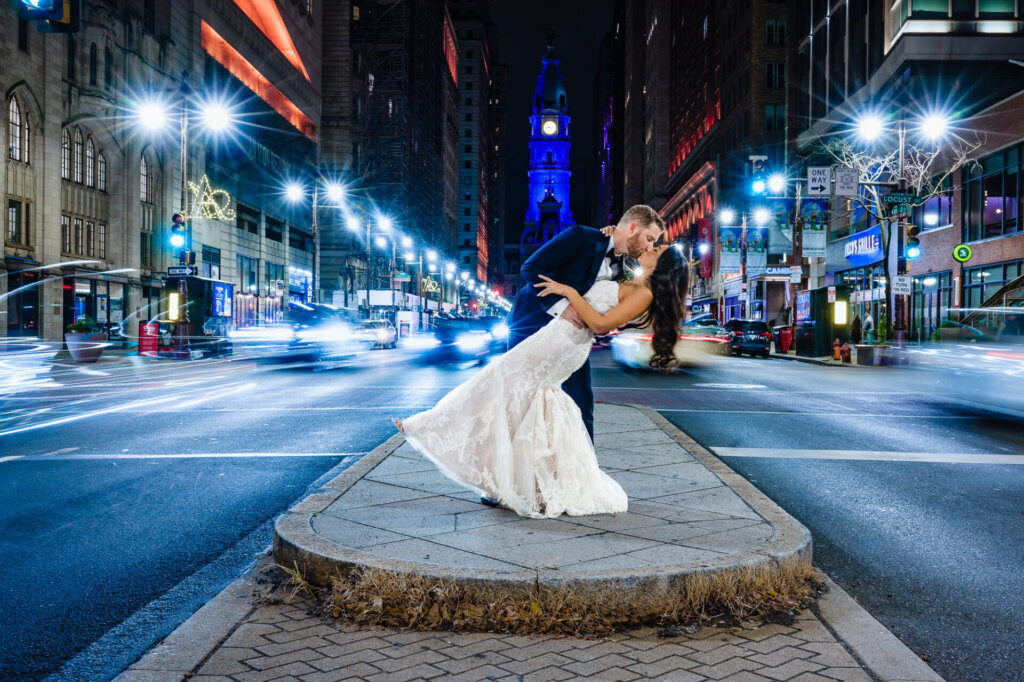 Bride and groom share a romantic dance dip on a city street at night, surrounded by vibrant lights, with blurred car trails and a beautifully illuminated cityscape in the background.