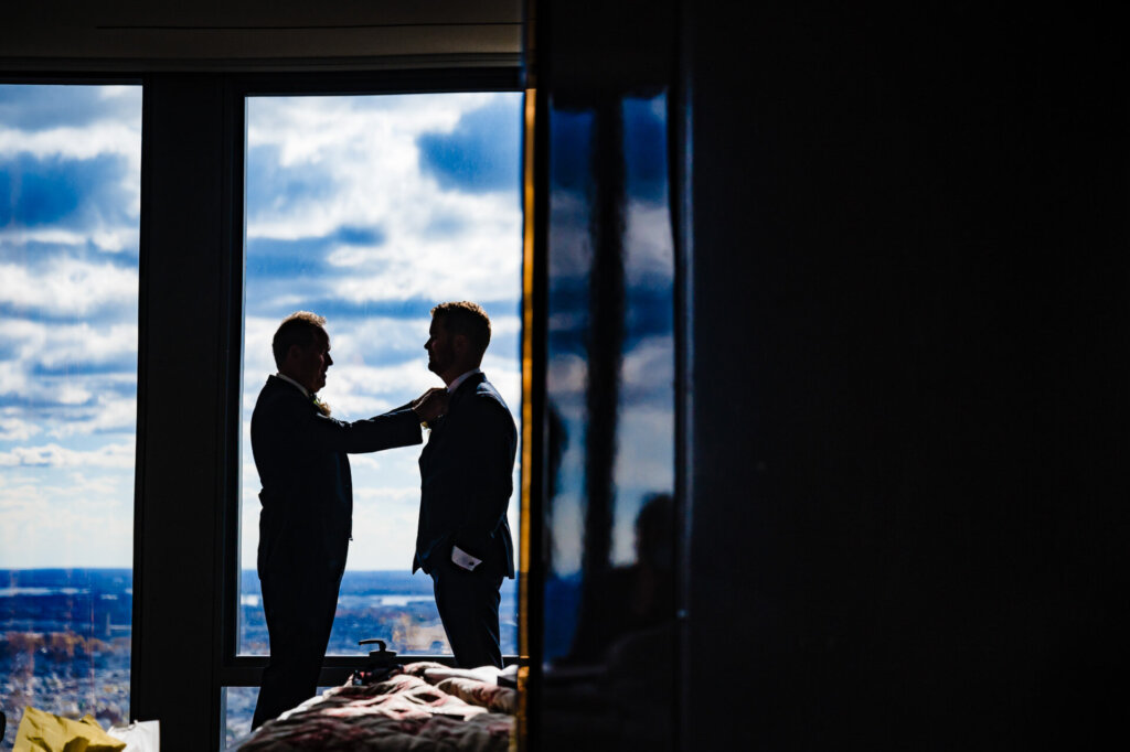 Silhouette of the groom and a groomsman adjusting attire by a window, overlooking the city skyline.