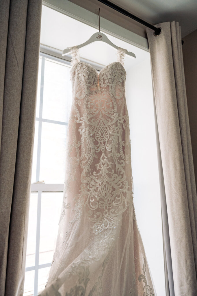 Wedding dress with intricate lace details hanging in front of a large window framed by beige curtains, softly illuminated by natural light.