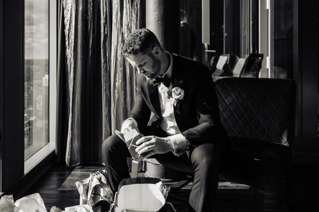 Groom sitting and carefully unwrapping a gift box, dressed in a black tuxedo with a white boutonniere.