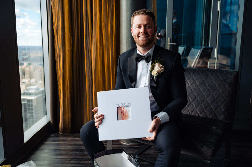 Groom holding up a framed photo or artwork, smiling warmly in his tuxedo.
