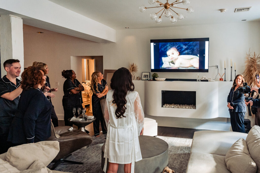 Bride standing in a modern living room with her bridal party and family members gathering around, with a TV and fireplace in the background.