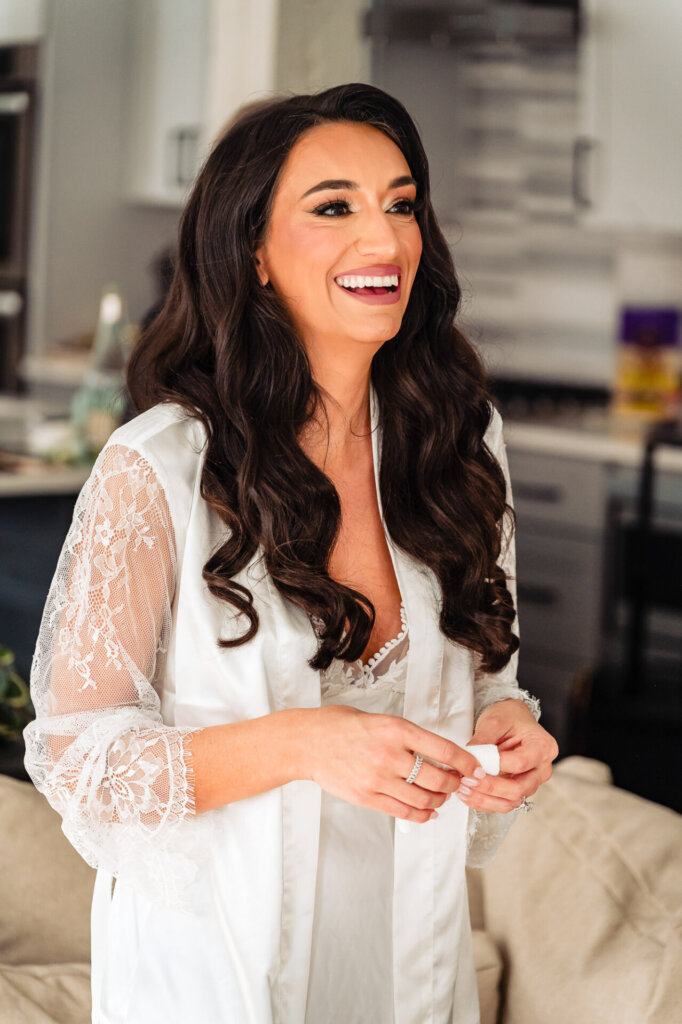 Bride smiling brightly while wearing a lace bridal robe, standing in a bright and elegant room during her wedding preparations.