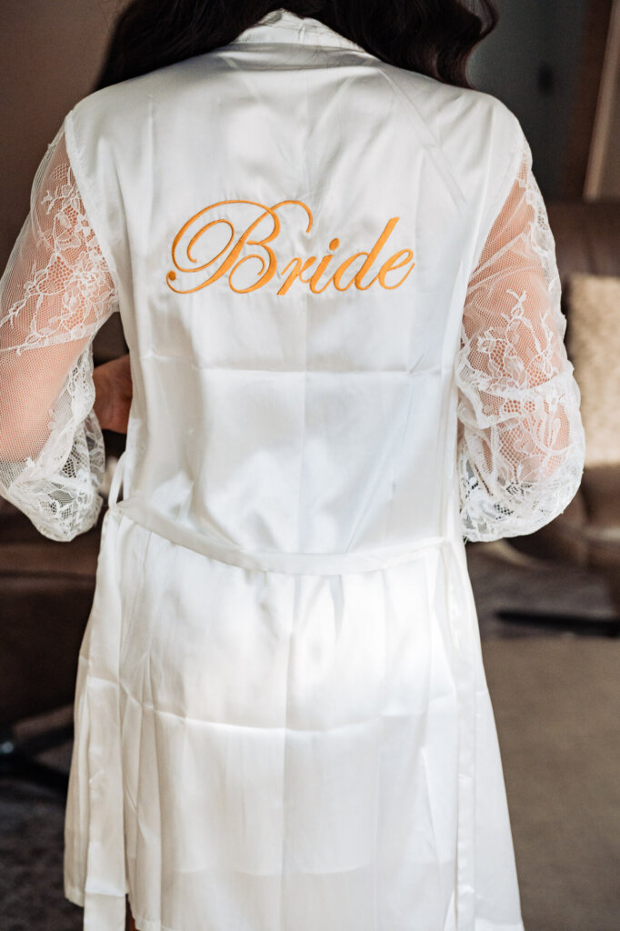 Back view of the bride wearing a white lace robe embroidered with the word 'Bride' in gold lettering, standing in a softly lit room.