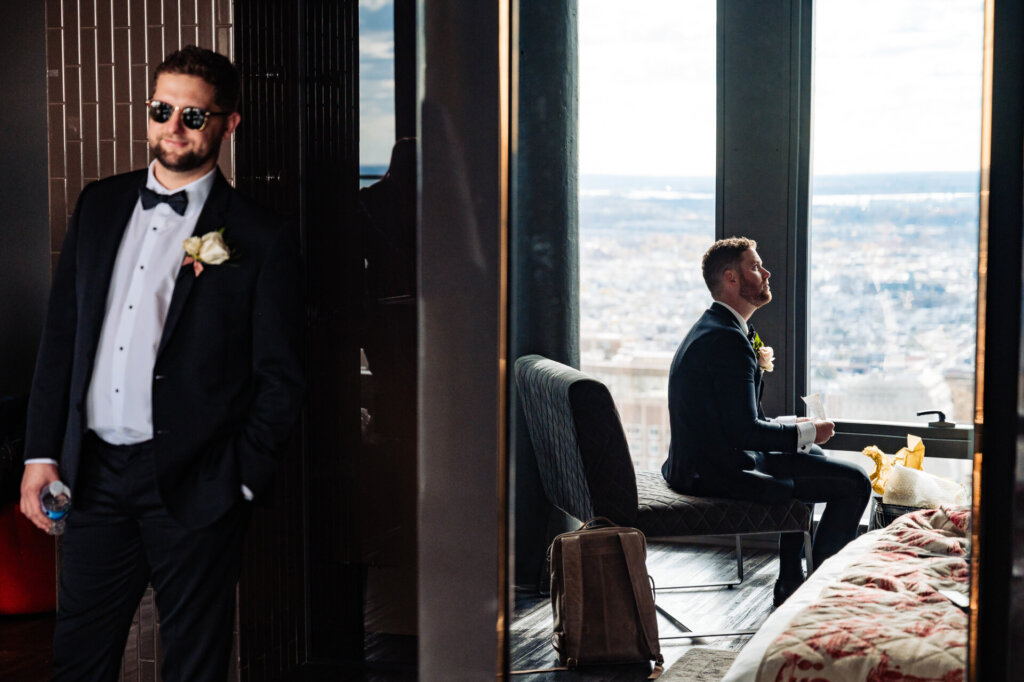 Groom sitting in a hotel suite, looking out at the expansive city skyline while preparing for the wedding with his groomsman in sunglasses, dressed in his wedding tuxedo, leaning casually against a wall while holding a bottle of water