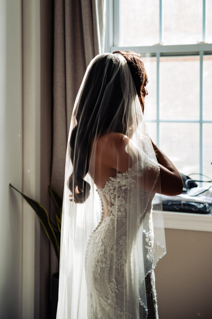 Bride standing by a large window, softly lit, with her veil and fitted lace gown elegantly framed by natural light.