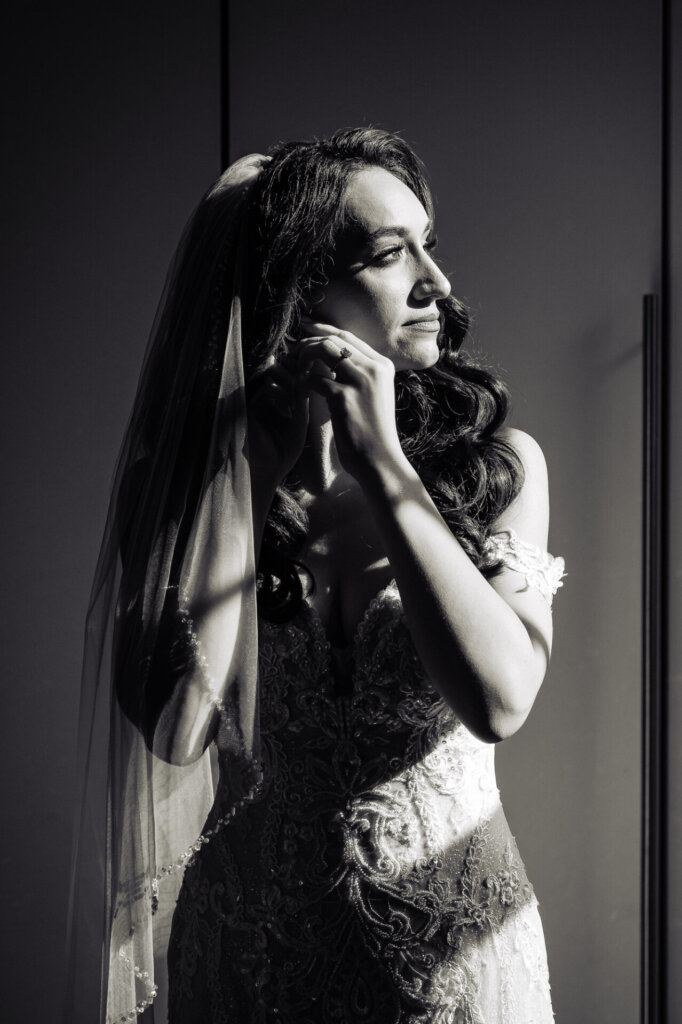 Bride, illuminated by sunlight, gazing thoughtfully out of the window, her lace gown and veil creating a delicate silhouette