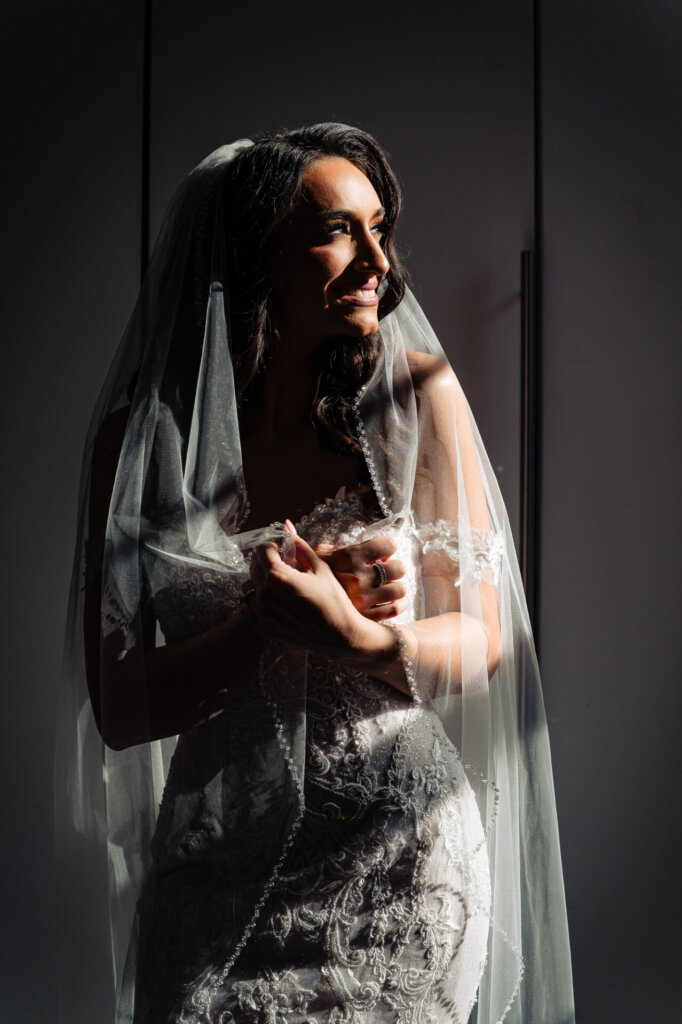 Bride holding her hands together, bathed in light and shadow, showcasing the intricate details of her lace gown.