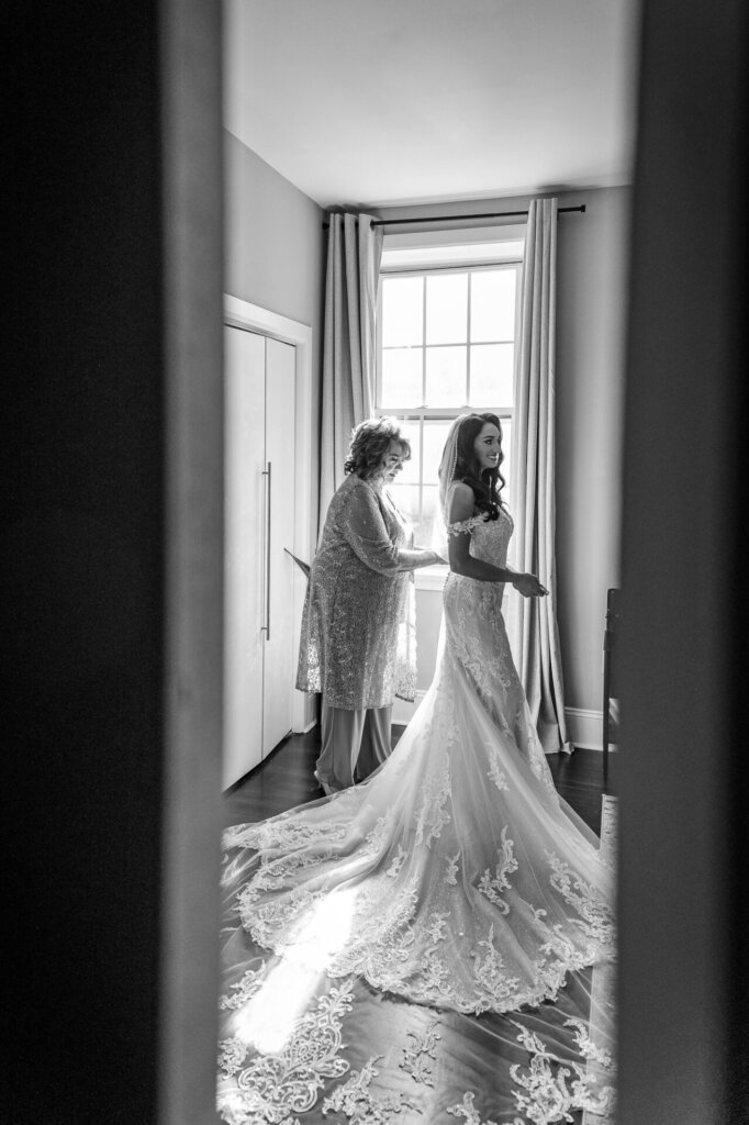 Black and white photo of the bride in a lace wedding gown with a long train, standing near a window as an older woman, possibly her mother, helps adjust her dress. The scene captures a candid and emotional moment of wedding preparation.