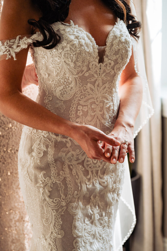 Close-up of the bride adjusting her lace wedding gown, showcasing the intricate detailing of the dress and her hands adorned with simple jewelry.