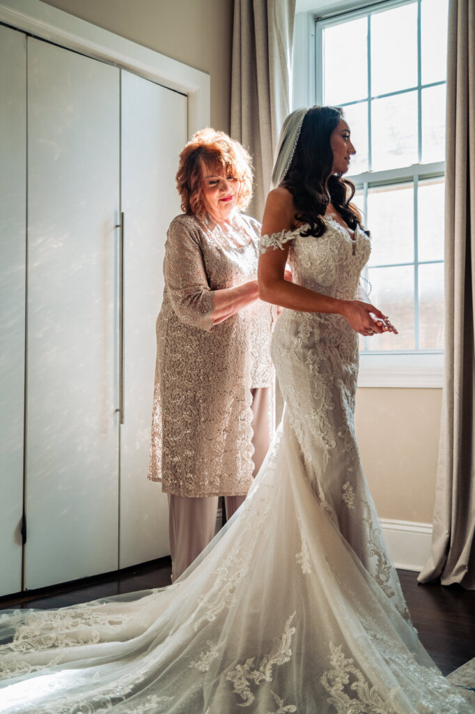 Bride in an off-the-shoulder lace wedding dress, standing still as her gown is adjusted by her mother wearing a glittering outfit, with natural light streaming in from the window.