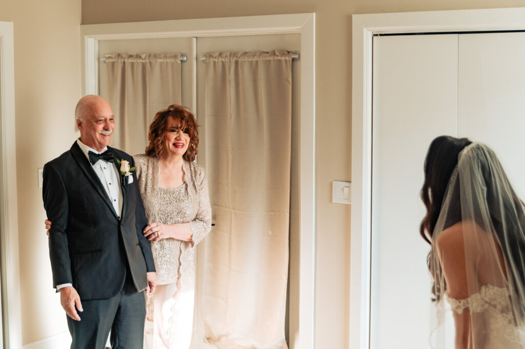 Father of the bride, dressed in a formal tuxedo, and the mother of the bride standing proudly as they admire their daughter in her wedding gown.