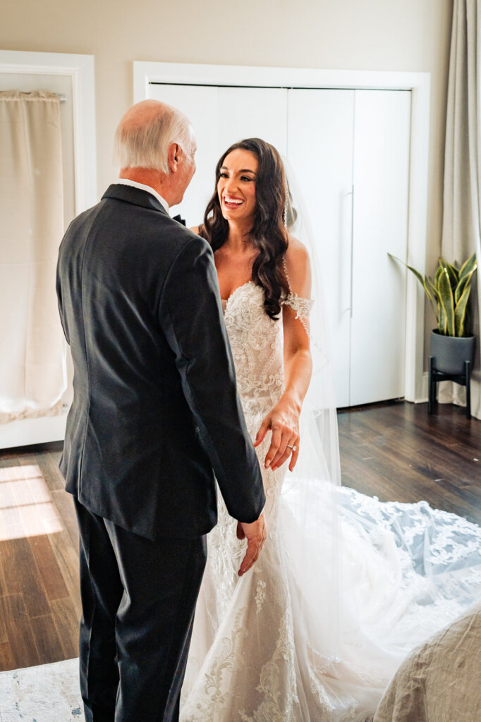 Father of the bride walking toward his daughter, who stands in her lace wedding gown, smiling as he reaches out for a warm embrace