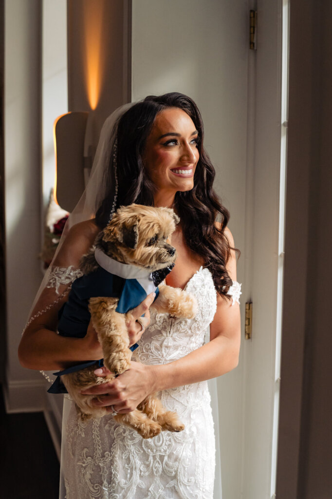 Bride holding her small dog, dressed in a formal tuxedo, as she smiles warmly in her lace wedding gown and veil.