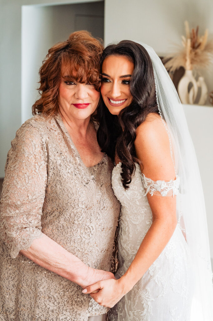 Close-up of the bride and her mother, with the mother wearing a glittering dress and the bride in a lace gown and veil, smiling while standing close together.