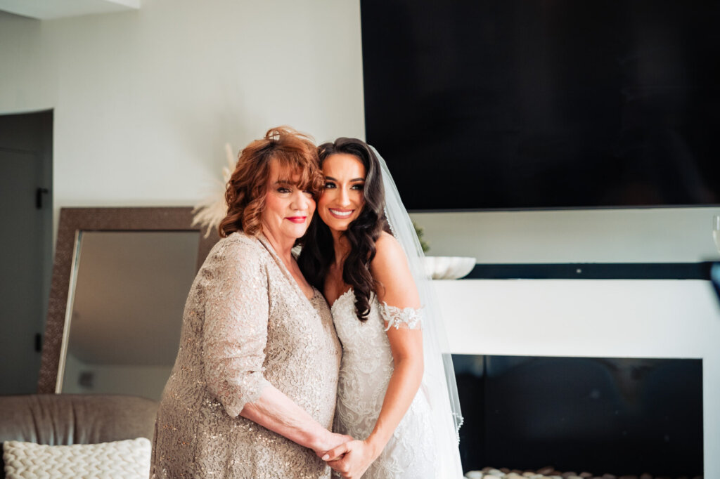 Bride and her mother standing side by side in a modern living room, holding hands and sharing a joyful moment before the wedding.