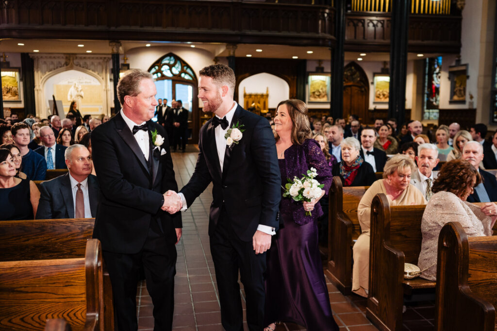 Father of the bride shaking hands with the groom at the altar, as the bride and her mother look on with warm smiles.