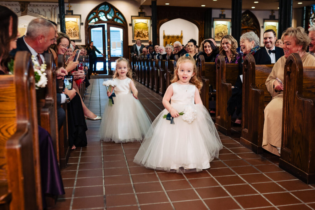 Two flower girls in white dresses walking down the aisle, holding small bouquets as they approach the altar.