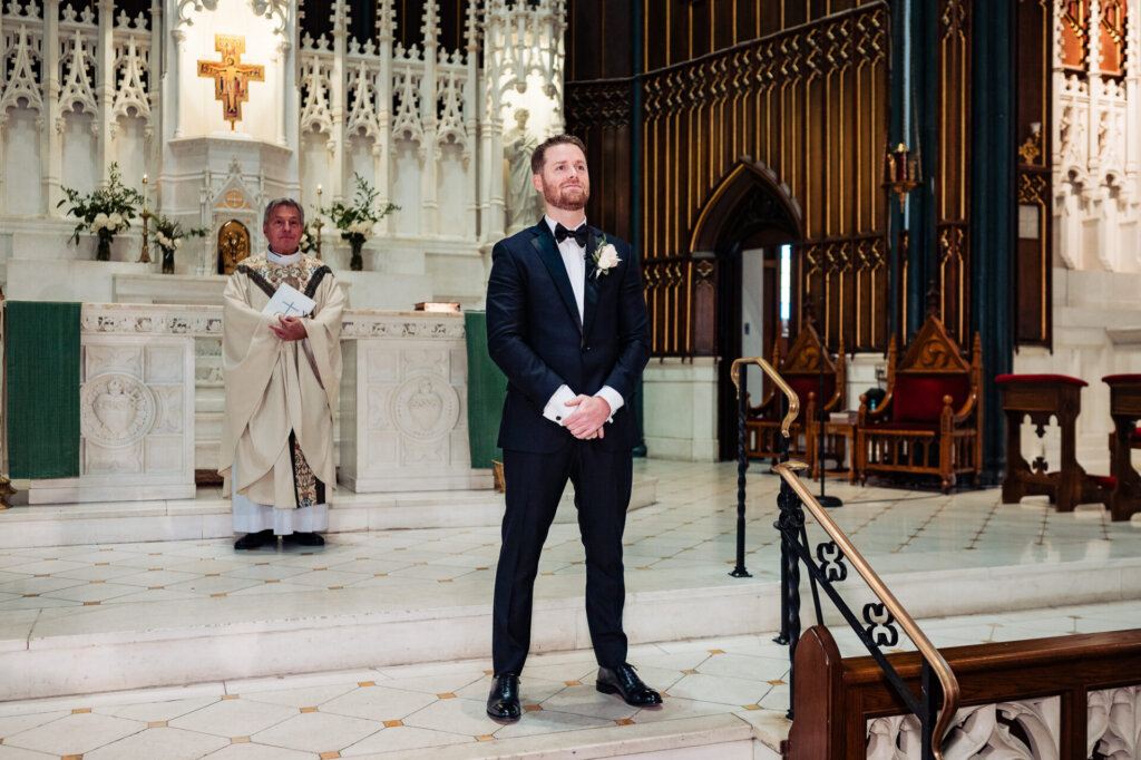 Groom standing at the altar in a grand cathedral, wearing a black tuxedo and boutonniere, waiting for the bride with an excited expression.