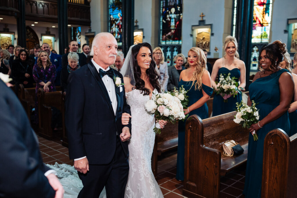 Bride walking down the aisle with her father, holding a bouquet of white and blush flowers, surrounded by guests in a cathedral.
