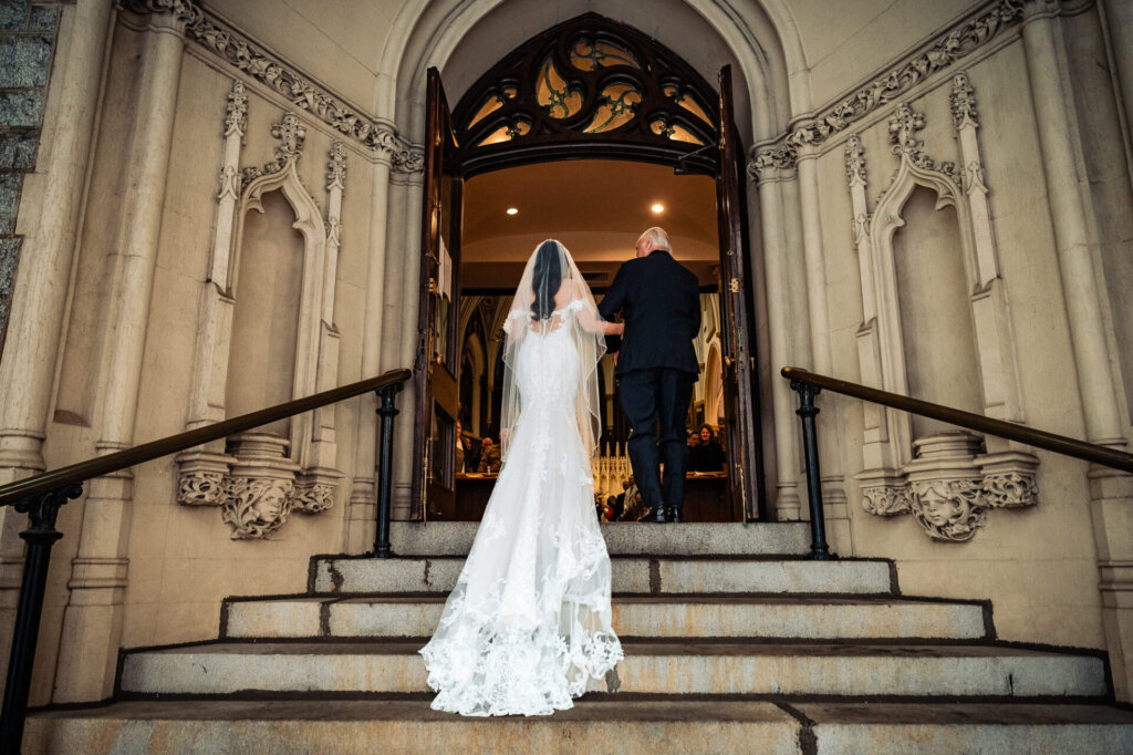 Bride walking up the stone steps of a historic church with her father, her long lace train cascading behind her as they approach the open doors.