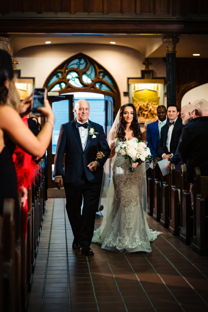 Bride and her father entering the church, her lace gown and veil glowing softly in the light as guests watch.