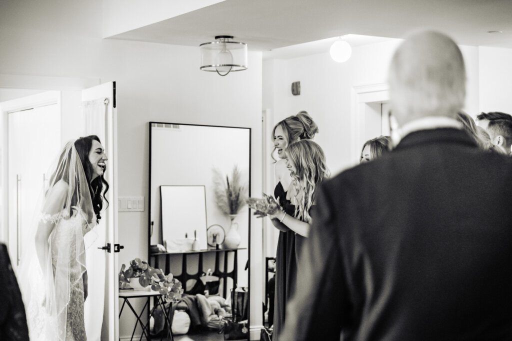 Black and white photo of the bride laughing and excitedly interacting with her bridal party as they prepare for the wedding, with her father visible from the back in the foreground.