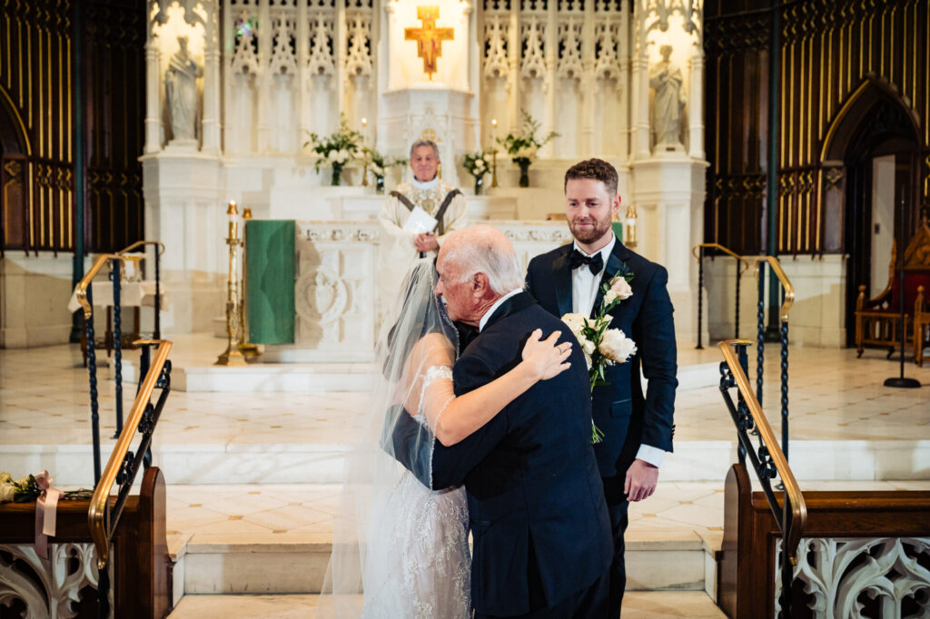 Father of the bride hugging his daughter at the altar as the groom looks on, with the grand cathedral altar in the background