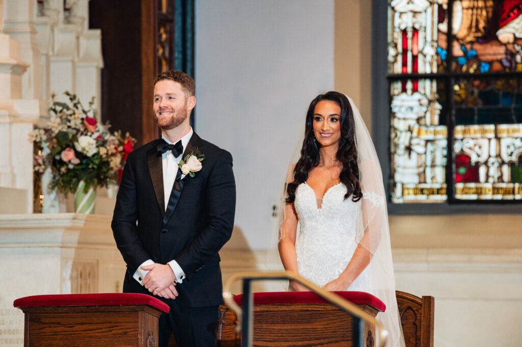 Bride standing at the altar with a calm smile, wearing her veil and intricate lace gown in front of the church’s ornate backdrop.