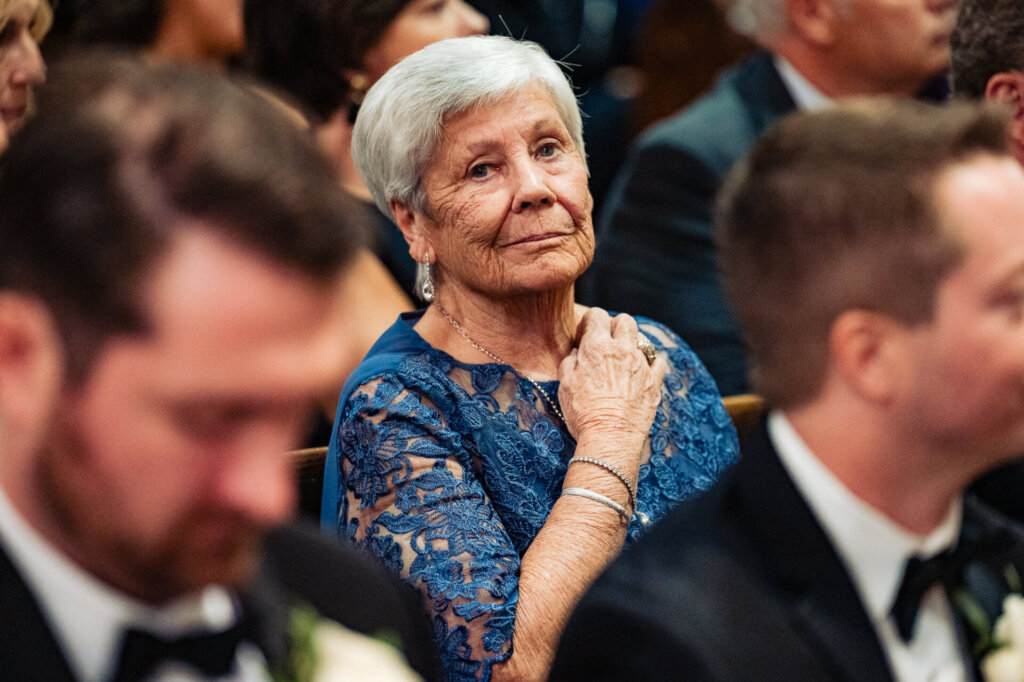 Grandmother of the bride, wearing a blue dress, clasping her hands to her chest with an emotional expression as she watches the ceremony.