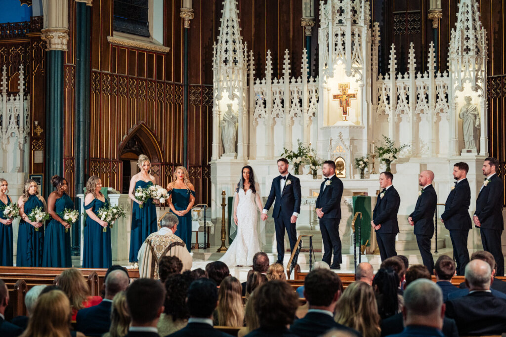 Wide-angle view of the bridal party and groomsmen standing at the altar, the ornate cathedral backdrop highlighting the ceremony setting