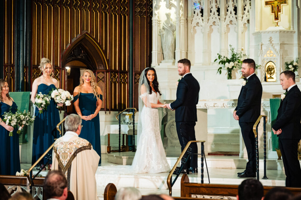 Bride smiling while holding the groom’s hands during the vows, as the bridal party looks on with joyful expressions on their A Classic Philadelphia Wedding