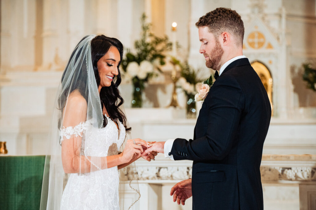 Bride placing a ring on the groom’s finger during their wedding ceremony at the altar, both smiling softly.
