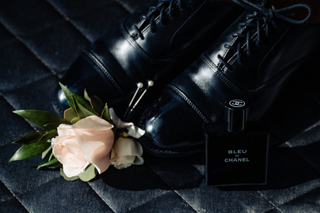 Black leather dress shoes with a boutonniere of pale pink roses and greenery, paired with a bottle of cologne on a dark surface.