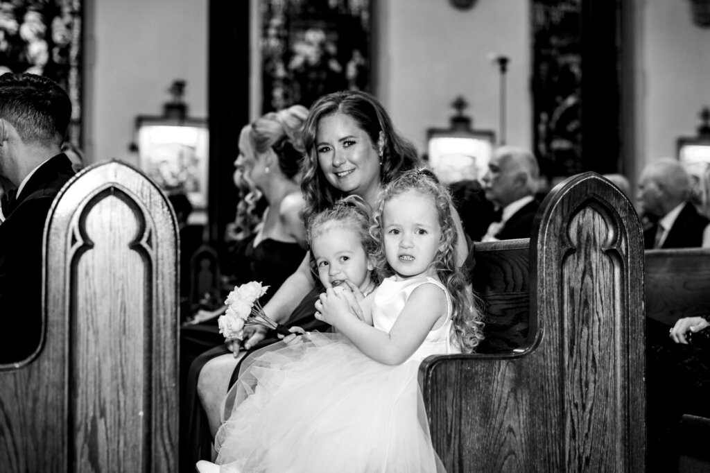 Bride’s guest sitting in the church pew, holding two young children, one in her arms and the other leaning against her, captured in black and white