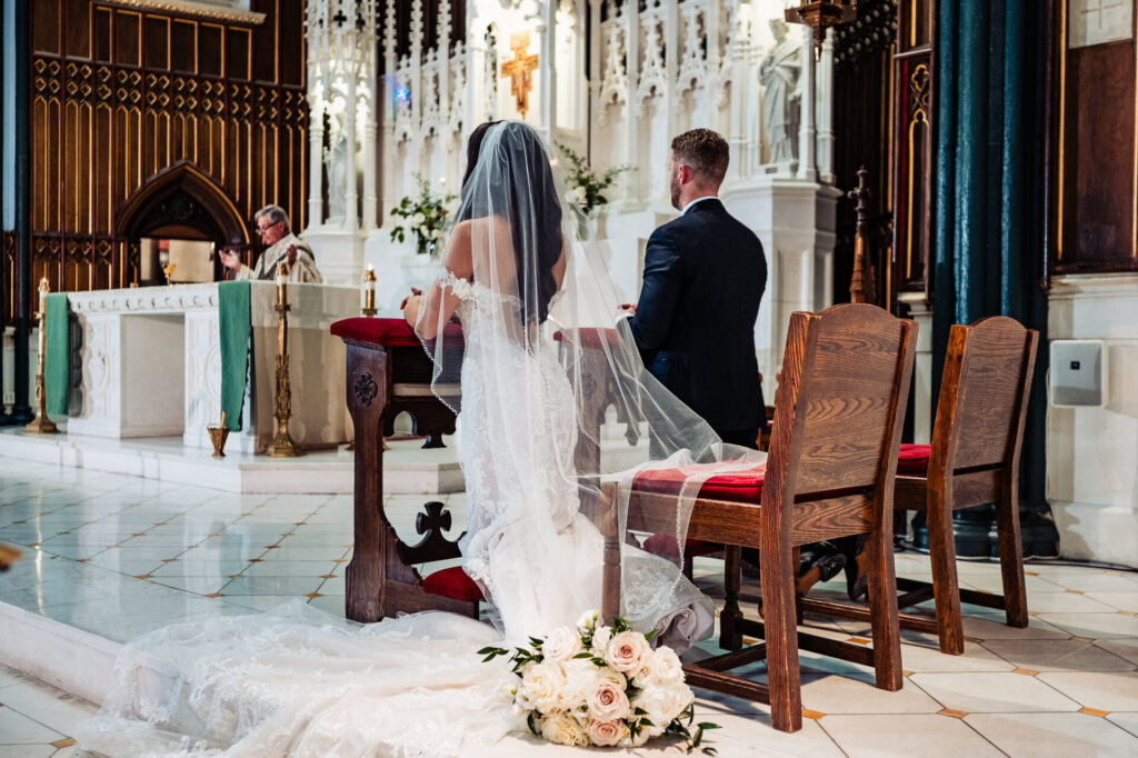 Bride and groom seated at the altar in a grand cathedral, with her bouquet placed nearby and the intricate architecture framing the scene.