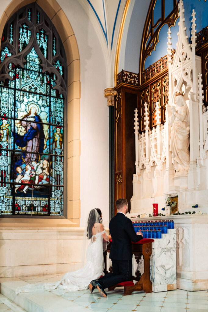 Bride and groom kneeling together in prayer at a side altar with a stained glass window depicting religious figures in the background, their hands clasped.