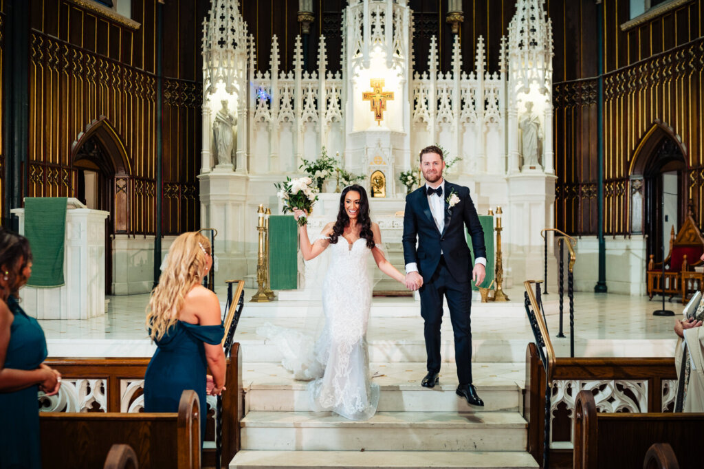 Bride and groom walking down the aisle as newlyweds, holding hands and smiling, while their bridal party looks on with joy.